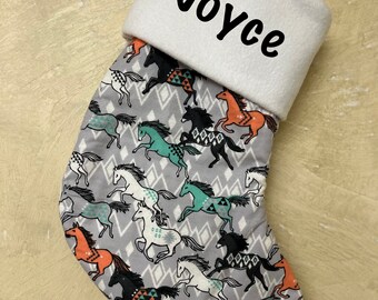 Może przedstawiać: Świąteczna skarpeta z białym mankietem i imieniem "Joyce" w czarnym skrypcie. Korpus skarpety zdobi wzór biegnących koni w różnych kolorach, w tym pomarańczowym, turkusowym i czarnym, na szarym tle w romby.