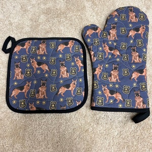 May include: A set of two blue oven mitts with a black border. The mitts feature a repeating pattern of German Shepherd dogs wearing police badges and gold stars.