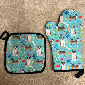 May include: A set of two blue and black oven mitts with a pattern of brown and white dogs playing with toys. The mitts are quilted and have a black trim.