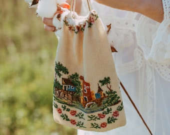 May include: A small, beige drawstring bag with a floral design and a scene of a house and a bridge embroidered on it. The bag is held by a person's hand.