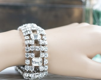 May include: A silver bracelet with a geometric design featuring multiple rows of clear rectangular and round rhinestones.