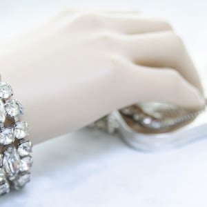 May include: A sparkling silver bracelet with clear, faceted rhinestones. The bracelet features a three-row design with both round and marquise-shaped stones. A silver clutch bag is in the background.
