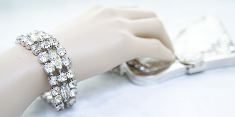 May include: A sparkling silver bracelet with clear, faceted rhinestones. The bracelet features a three-row design with both round and marquise-shaped stones. A silver clutch bag is in the background.