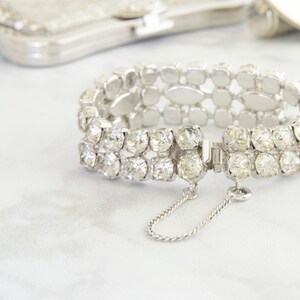 May include: A silver-toned bracelet with two rows of clear, sparkling rhinestones. The bracelet has a secure clasp with a safety chain. A silver mesh purse is in the background.