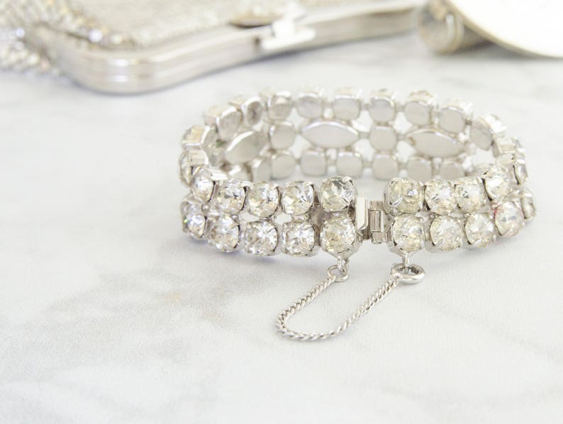 May include: A silver-toned bracelet with two rows of clear, sparkling rhinestones. The bracelet has a secure clasp with a safety chain. A silver mesh purse is in the background.