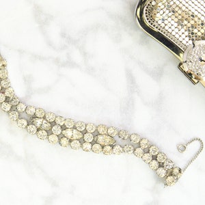 May include: A vintage rhinestone bracelet with a silver-tone metal setting. The bracelet features three rows of round and oval-shaped clear rhinestones. A silver mesh clutch with a rhinestone clasp is in the background.