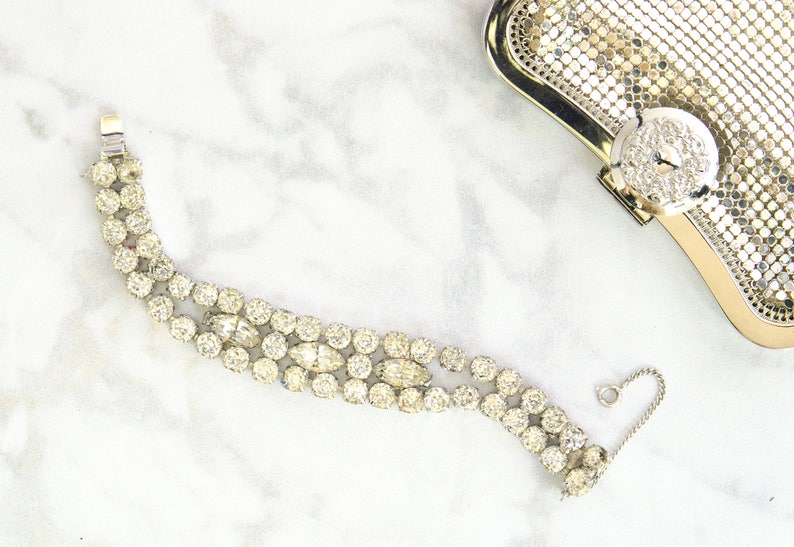 May include: A vintage rhinestone bracelet with a silver-tone metal setting. The bracelet features three rows of round and oval-shaped clear rhinestones. A silver mesh clutch with a rhinestone clasp is in the background.
