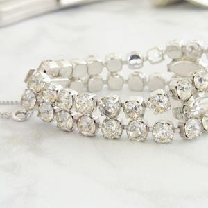 May include: A silver-toned bracelet with three rows of clear, sparkling rhinestones. The bracelet has a delicate chain and clasp closure. The stones are square and round, set in a silver metal. The jewelry is displayed on a white surface.