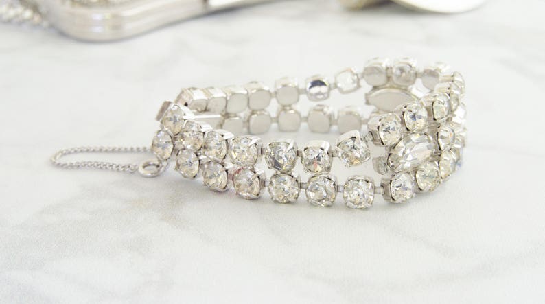 May include: A silver-toned bracelet with three rows of clear, sparkling rhinestones. The bracelet has a delicate chain and clasp closure. The stones are square and round, set in a silver metal. The jewelry is displayed on a white surface.