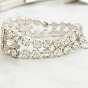 May include: A silver-toned bracelet with three rows of clear, sparkling rhinestones. The bracelet has a secure clasp and a safety chain. The stones are set in a square or rectangular shape, creating a glamorous accessory.