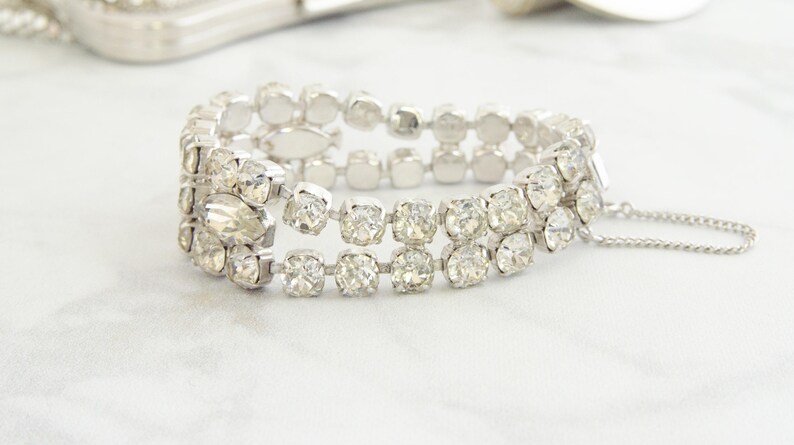 May include: A silver-toned bracelet with three rows of clear, sparkling rhinestones. The bracelet has a secure clasp and a safety chain. The stones are set in a square or rectangular shape, creating a glamorous accessory.