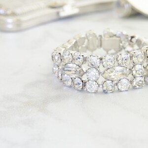 May include: A sparkling silver bracelet with clear, faceted crystals. The bracelet features a series of round and oval-shaped stones set in a silver metal band. The jewelry is displayed on a white surface, with a blurred silver accessory in the background.