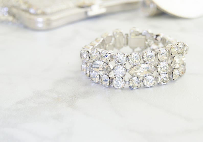 May include: A sparkling silver bracelet with clear, faceted crystals. The bracelet features a series of round and oval-shaped stones set in a silver metal band. The jewelry is displayed on a white surface, with a blurred silver accessory in the background.
