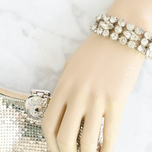 May include: A sparkling silver-toned bracelet with clear, round and marquise-cut rhinestones. The bracelet is worn on a hand, next to a silver mesh clutch with a rhinestone clasp. The jewelry is set against a white marble background.