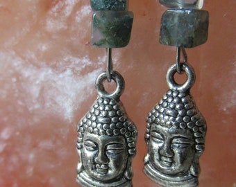 May include: A pair of silver-toned earrings featuring a Buddha head pendant. Each earring has three square, green-toned beads above the pendant. The background is a soft, pinkish-orange color.