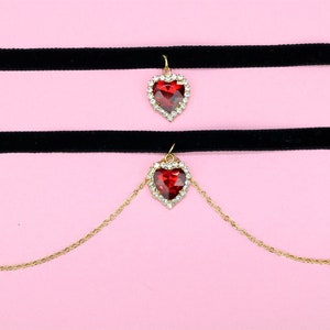 May include: A black velvet choker necklace with two red heart pendants. The top pendant is attached to the choker, while the bottom pendant hangs from a gold chain.