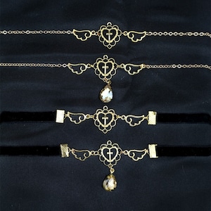 May include: Four gold necklaces with a heart and cross pendant and angel wings. The necklaces are layered on a black velvet background. The bottom two necklaces have a teardrop-shaped crystal hanging from the pendant.