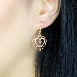 May include: Gold-tone heart-shaped earrings with a cross design. The earrings are dangling and have a delicate, intricate design.