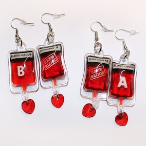 Puede incluir: Un par de pendientes con colgantes de bolsa de sangre roja con las palabras "Blood Group" y "Safe Blood" impresas en ellos. Las bolsas de sangre están llenas de líquido rojo y tienen un tubo de plástico transparente que cuelga de la parte inferior. Cada colgante está unido a un cristal rojo en forma de corazón.