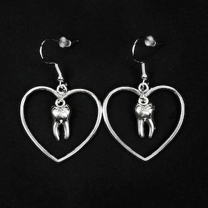 May include: Silver heart-shaped earrings with a silver tooth charm hanging from each heart.