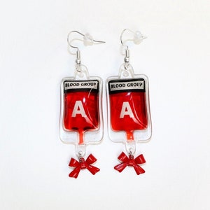 May include: A pair of earrings shaped like blood bags with red liquid inside. Each bag has a white label that says "BLOOD GROUP" and a red letter "A". A red bow hangs from the bottom of each bag.