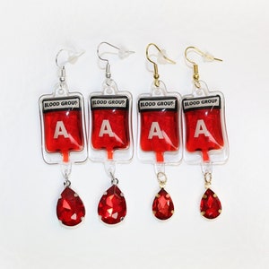 May include: Four pairs of earrings featuring a blood bag charm and a red teardrop crystal. The blood bags are clear and filled with red liquid. The bags have the text "Blood Group A" printed on them.