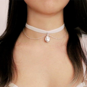 May include: A white velvet choker necklace featuring a silver chain and a teardrop-shaped crystal pendant. The necklace is worn around the neck, with the pendant suspended in the centre. A delicate piece of jewellery.