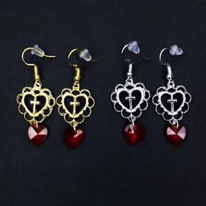 May include: Four pairs of dangle earrings with a heart-shaped design. Each earring features a filigree heart with a cross inside and a dangling red heart-shaped crystal.