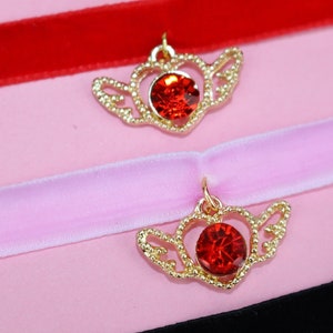 May include: Two gold heart-shaped chokers with red crystal accents and delicate wings. The chokers are displayed on a pink background.