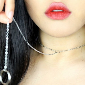 May include: A silver chain necklace with a ring pendant. The chain is layered with a shorter chain that has a small circle charm.