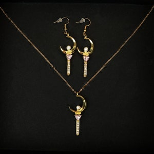 May include: A gold necklace and earrings set featuring a crescent moon and a pink heart design. The pendant and earrings are adorned with sparkling rhinestones.
