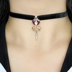 Puede incluir: Un collar gargantilla de terciopelo negro con un colgante de corazón dorado con un centro de esmalte rojo y diamantes de imitación transparentes. El colgante también tiene un lazo dorado y un tallo dorado largo y delgado.