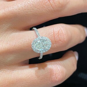 May include: A diamond ring with a large oval-cut center stone surrounded by a halo of smaller diamonds. The ring is set on a delicate band, and the diamonds sparkle brightly. The ring is on a person's finger.