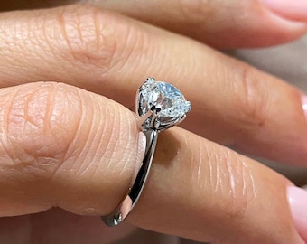 May include: A silver engagement ring with a large, clear, heart-shaped gemstone. The ring is on a finger with a light pink nail polish. The background is a soft, neutral color.