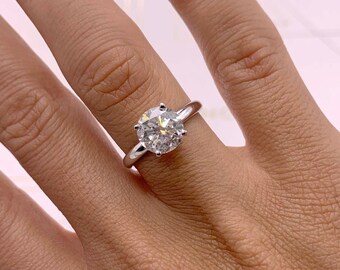 May include: A silver engagement ring with a large, round-cut diamond. The ring has a classic solitaire design with a thin band. The diamond sparkles brightly. The ring is shown on a person's hand.