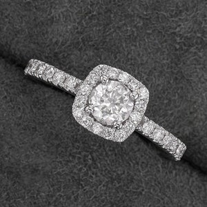 Pode incluir: Um anel de diamante com uma crava&ccedil;&atilde;o de halo quadrado. A pedra central &eacute; redonda, rodeada por diamantes menores. A banda tamb&eacute;m &eacute; incrustada com diamantes, criando um efeito brilhante. O anel est&aacute; sobre uma superf&iacute;cie cinza escuro.
