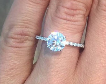 Könnte beinhalten: Eine Hand mit manikürten Nägeln, die einen Diamant-Verlobungsring zeigt. Der Ring hat einen großen, rund geschliffenen Diamanten als Mittelstein und ein zartes Band mit kleineren Akzentdiamanten. Die Nägel haben eine natürliche rosa Basis mit weißen Spitzen.