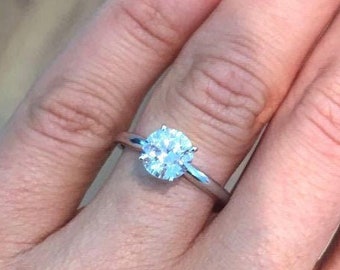 May include: A solitaire diamond ring with a round-cut diamond set in a thin, silver band. The ring is shown on a person's finger, with a close-up view of the sparkling gemstone.