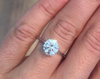 May include: A silver engagement ring with a large, round, clear gemstone. The ring is set on a hand with manicured nails. The nails are painted with a light pink polish and white tips.