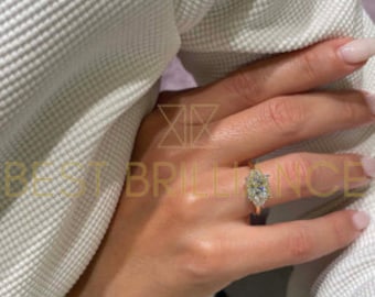 May include: A gold ring with a large, oval-shaped diamond center stone, surrounded by smaller diamonds. The ring is on a person's finger, with a white sweater in the background. The text "BEST BRILLIANCE" is visible.