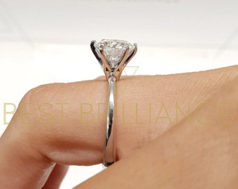 May include: A silver engagement ring with a large, clear, round-cut diamond. The diamond is held in place by four prongs. The ring is on a finger, with the words "BEST BRILLIANCE" visible in the background.