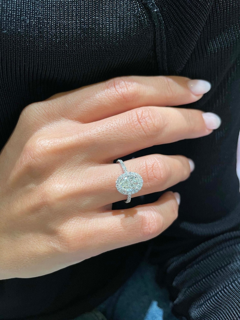May include: A diamond ring with a large oval-cut center stone surrounded by a halo of smaller diamonds. The ring is set on a delicate band, and the diamonds sparkle brightly. The ring is on a person's finger.