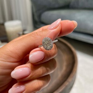 May include: A silver ring with a large oval-cut diamond center stone, surrounded by a halo of smaller diamonds. The ring's band also features small diamonds. The ring is held up against a blurred background.