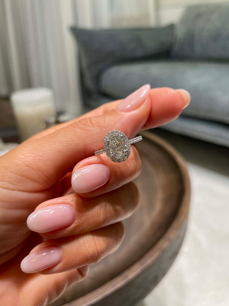 May include: A silver ring with a large oval-cut diamond center stone, surrounded by a halo of smaller diamonds. The ring's band also features small diamonds. The ring is held up against a blurred background.