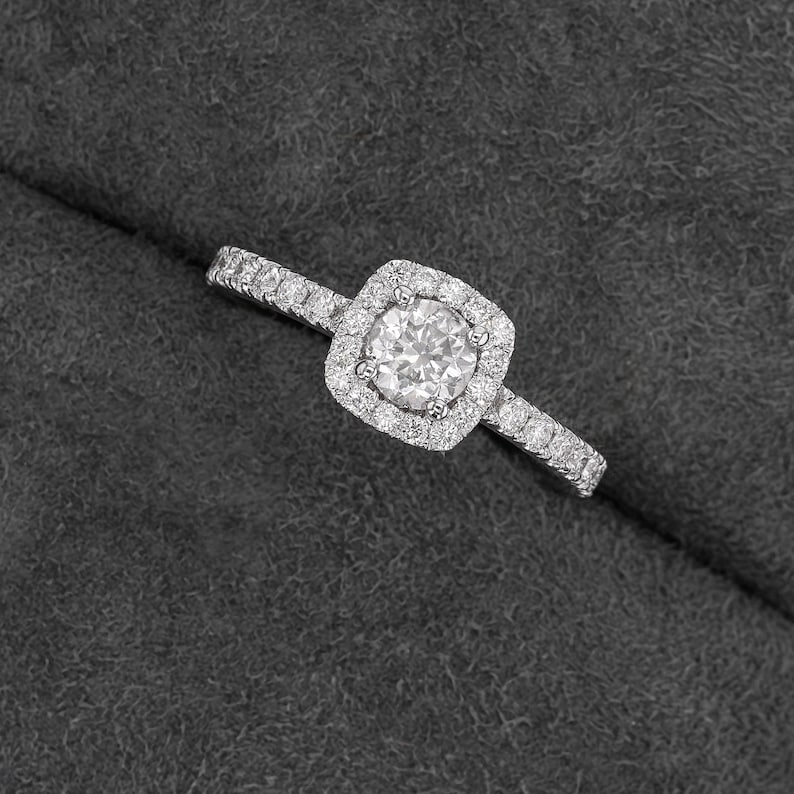 Pode incluir: Um anel de diamante com uma crava&ccedil;&atilde;o de halo quadrado. A pedra central &eacute; redonda, rodeada por diamantes menores. A banda tamb&eacute;m &eacute; incrustada com diamantes, criando um efeito brilhante. O anel est&aacute; sobre uma superf&iacute;cie cinza escuro.