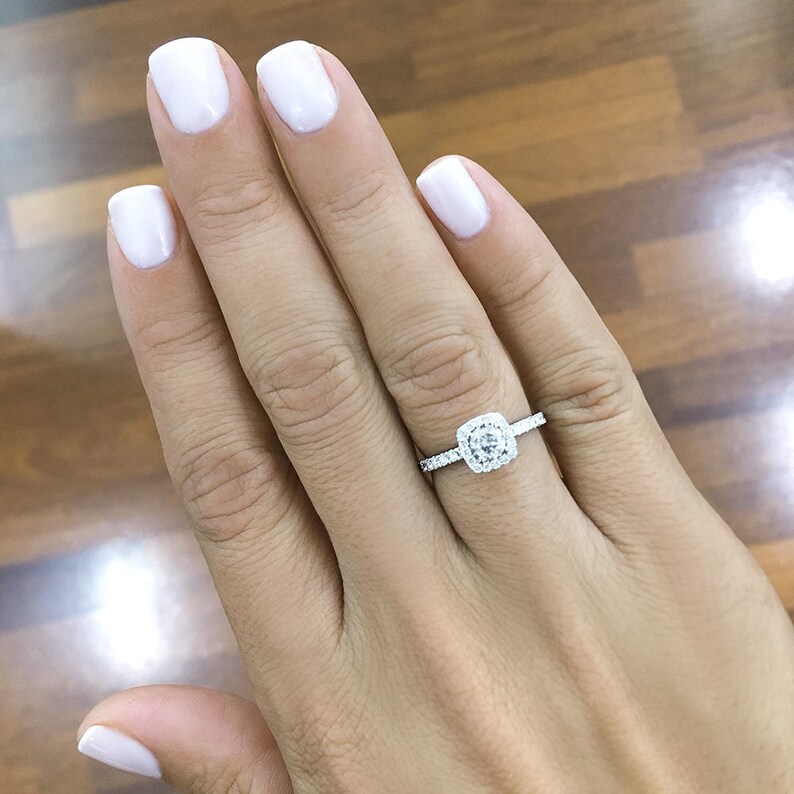 Pode incluir: Um anel de noivado de diamantes com uma crava&ccedil;&atilde;o de halo quadrado. O anel apresenta um diamante central de corte quadrado rodeado por diamantes menores. A banda tamb&eacute;m &eacute; adornada com pequenos diamantes. O anel est&aacute; no dedo de uma pessoa.