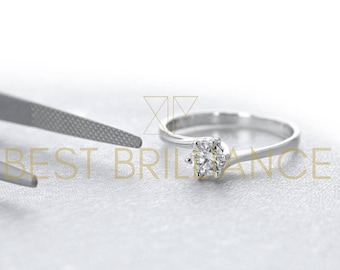 Puede incluir: Primer plano de un anillo de compromiso plateado con un diamante transparente. El anillo está sobre un fondo blanco, con una herramienta en el fondo. El texto "BEST BRILLIANCE" es visible.