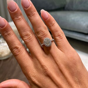 May include: A diamond engagement ring with a large oval-cut center stone surrounded by a halo of smaller diamonds. The ring is set on a delicate, diamond-studded band. The hand has long, manicured nails.
