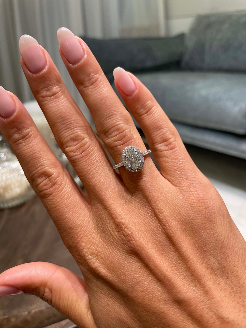May include: A diamond engagement ring with a large oval-cut center stone surrounded by a halo of smaller diamonds. The ring is set on a delicate, diamond-studded band. The hand has long, manicured nails.