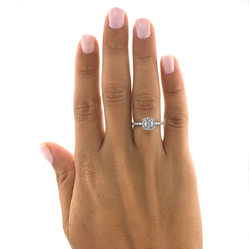 Pode incluir: Um anel de prata com um halo quadrado de pequenas pedras que circundam uma pedra central maior, de corte quadrado. O anel &eacute; exibido em uma m&atilde;o com unhas cuidadas.